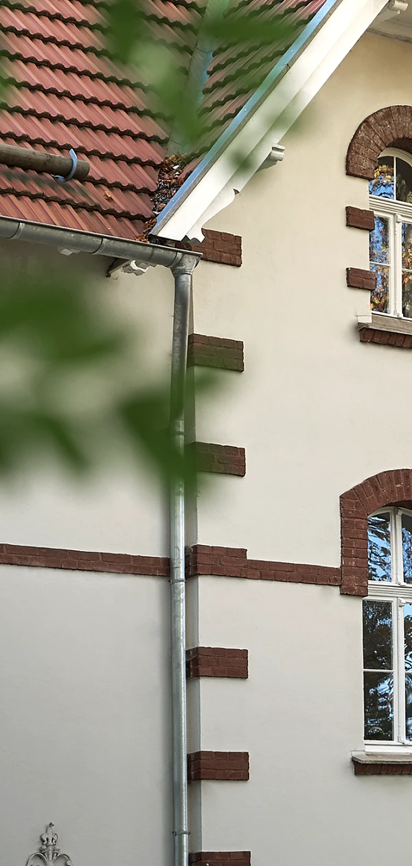 Vorderansicht eines Altbaus mit roten Ziegeln und Rundfenstern, durch Blätter gerahmt