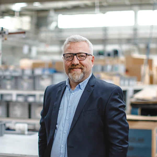 Mann mit Brille und grauem Bart in Produktionshalle, freundlich lächelnd